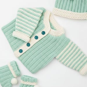 May include: A mint green crochet baby set, including a cardigan with teal buttons, a beanie with white pom-poms, and matching booties. The set features white striped accents and a cream-coloured trim, ideal for a newborn.