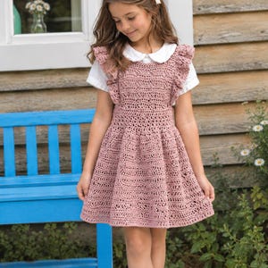 May include: A dusty rose crochet pinafore dress with ruffled shoulder straps and a textured, open-work pattern. The dress is worn over a short-sleeved white shirt with a rounded collar. A small white hair bow is clipped to the side of the hair.