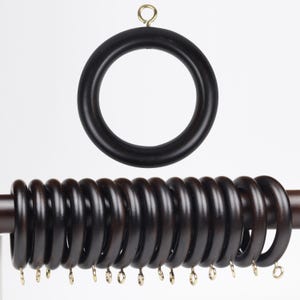 May include: Dark brown wooden curtain rings with brass-colored hooks. The rings are circular and have a smooth finish. One ring is shown separately, while the others are attached to a rod. The background is plain white.
