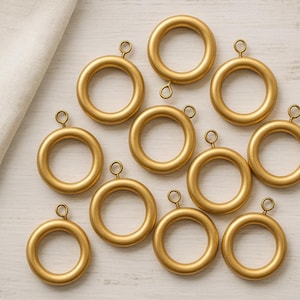 Op de afbeelding: Een verzameling goudkleurige gordijnringen met kleine oogjes. De ringen zijn gerangschikt op een lichtgekleurd houten oppervlak, met een gevouwen witte doek in de linkerbovenhoek. De ringen zijn cirkelvormig en hebben een matte afwerking.