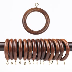May include: Wooden curtain rings in a dark brown hue. The rings are circular with a smooth finish and small, gold-coloured hooks. One ring is shown separately above the others. The rings are designed for hanging curtains.