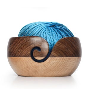 May include: A two-toned wooden yarn bowl with a light blue ball of yarn inside. The bowl is made of light and dark wood, with a swirl-shaped yarn guide. The yarn is a vibrant shade of blue.
