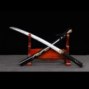 Może przedstawiać: Japoński miecz katana ze srebrnym ostrzem i czarną rękojeścią, spoczywający na drewnianym stojaku. Czarna pochwa ma złote zdobienia. Miecz jest tradycyjną bronią.