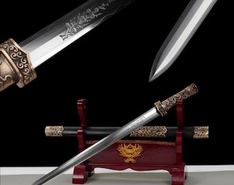 Puede incluir: Una espada decorativa con una hoja plateada y un mango de bronce adornado. La hoja presenta diseños florales grabados. La espada descansa sobre un soporte de madera rojo con una vaina negra, mostrando una estética tradicional.