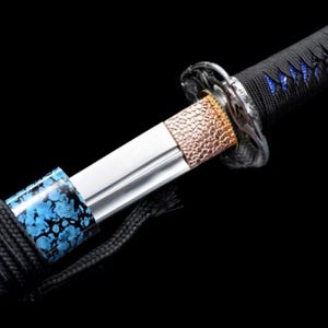 May include: Close-up of a Japanese katana sword. The blade is silver, with a copper-colored hilt and a black handle wrapped with black cord. The handle has blue accents. The sword is set against a black background.