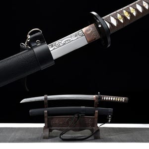 Può includere: Una spada katana giapponese con fodero e impugnatura in pelle nera. La lama è argentata con disegni incisi e l'impugnatura è avvolta in tessuto marrone. La spada poggia su un supporto di legno.