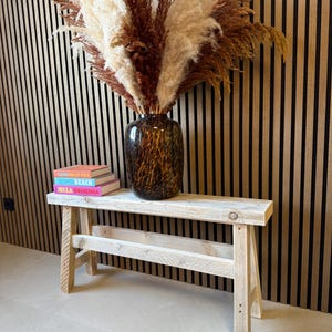 Könnte beinhalten: Eine rustikale Holzbank mit hellem Finish trägt eine dunkle, gemusterte Vase mit getrocknetem Pampasgras. Drei Bücher mit farbigen Buchrücken liegen auf der Bank. Der Hintergrund ist eine vertikale Holzpaneelwand.