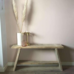 May include: A light-colored wooden bench with a lower shelf, set against a pale pink wall. A white vase with tall, feathery dried plants, a candle, and a small glass jar are on the bench. A black and white patterned rug is in the foreground.