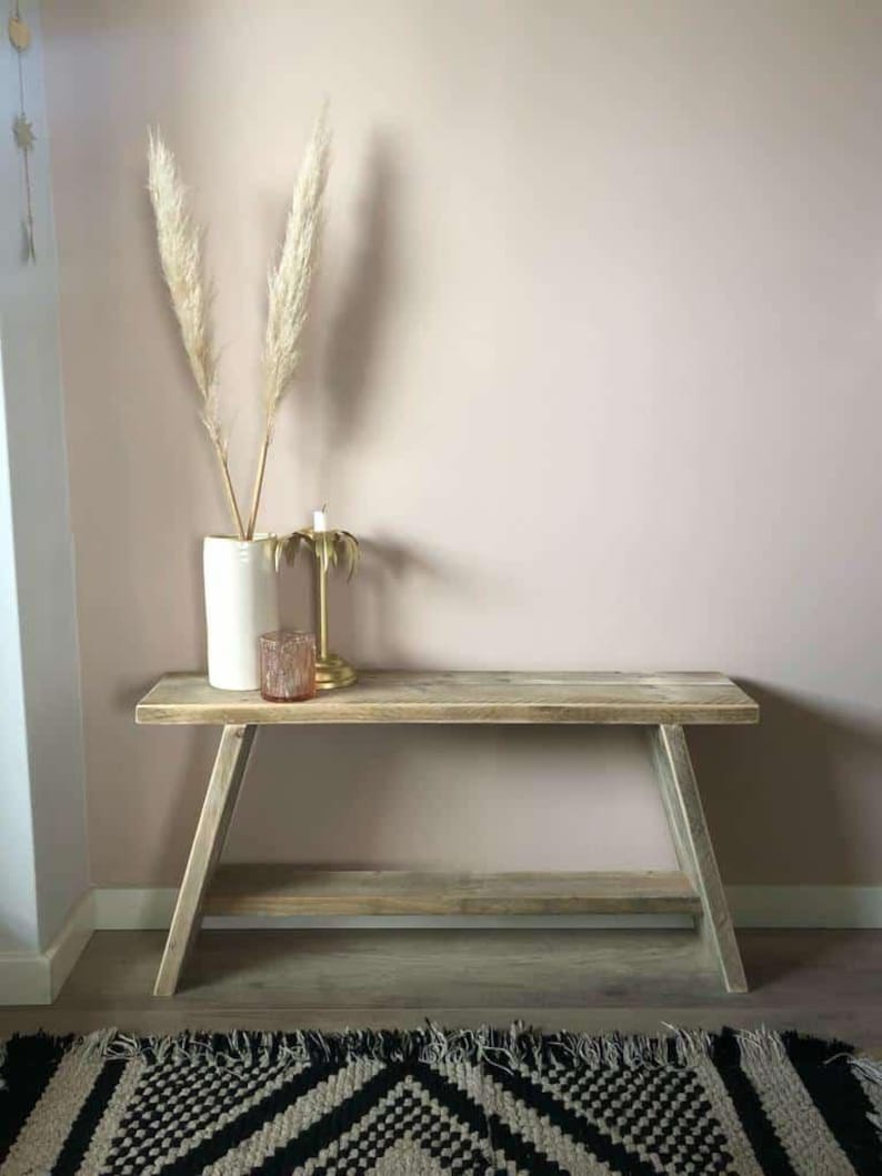 May include: A light-colored wooden bench with a lower shelf, set against a pale pink wall. A white vase with tall, feathery dried plants, a candle, and a small glass jar are on the bench. A black and white patterned rug is in the foreground.