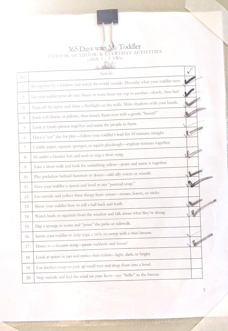 May include: A white paper document titled 365 Days with My Toddler: Indoor, Outdoor, & Everyday Activities (Age 1-3 Yrs) held by a black binder clip. The page features a numbered list of twenty activities with handwritten checkmarks next to most items.