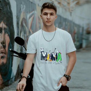 May include: White t-shirt with a colorful graphic featuring the word "MAMA" with Halloween-themed characters. The text "This MAMA loves her little Nightmares" is below the word. The shirt is worn by a person.