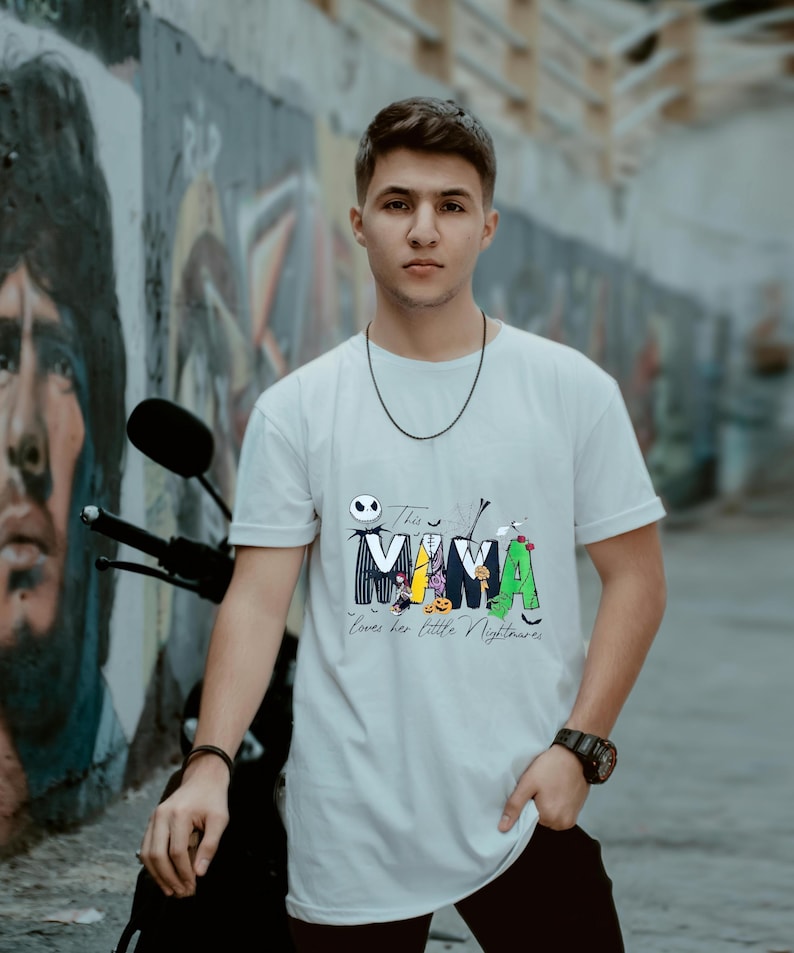 May include: White t-shirt with a colorful graphic featuring the word "MAMA" with Halloween-themed characters. The text "This MAMA loves her little Nightmares" is below the word. The shirt is worn by a person.