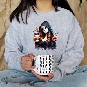 May include: A gray sweatshirt featuring a graphic of a woman in a hockey mask holding a coffee cup and making a peace sign. The sweatshirt is paired with a white mug with black vertical dashes.