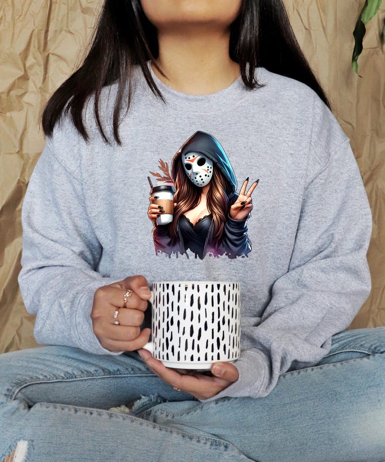 May include: A gray sweatshirt featuring a graphic of a woman in a hockey mask holding a coffee cup and making a peace sign. The sweatshirt is paired with a white mug with black vertical dashes.
