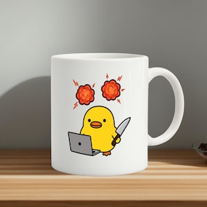 May include: White ceramic mug featuring a cartoon yellow chick with a knife and a laptop. The chick is positioned in front of two orange explosion graphics. The mug has a curved handle and is set on a wooden surface.