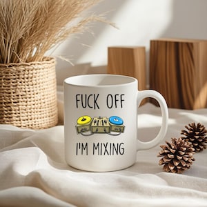 May include: White ceramic mug with the text "FUCK OFF I'M MIXING" and a cartoon illustration of DJ turntables. The turntables have yellow and blue record players. The mug is on a white cloth.