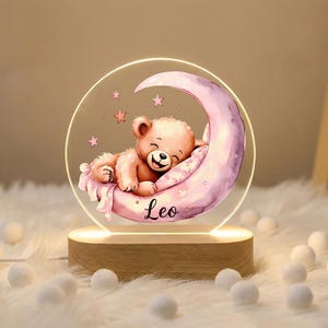 May include: A night light featuring a sleeping teddy bear on a crescent moon. The bear is light brown, resting on a pink moon with stars. The name "Leo" is written below. The light sits on a wooden base.