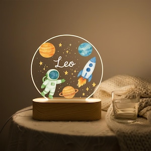 以下が含まれることがあります： 宇宙をテーマにした、丸い形の照明付きナイトライト。宇宙飛行士、ロケット、惑星、星が描かれています。「Leo」という名前が筆記体で書かれています。ベースは長方形の木製ブロックです。