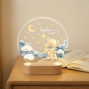 May include: A personalized night light featuring a teddy bear holding a star balloon, set against a backdrop of clouds, stars, and a crescent moon. The name "Sophia" and the date "09.15.2024" are displayed. The light has a wooden base.