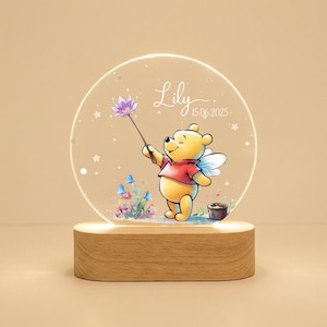 Puede incluir: Una luz de noche de Winnie the Pooh con una base de madera. El círculo acrílico transparente presenta un dibujo animado de Winnie the Pooh sosteniendo una flor, con el nombre "Lily" y la fecha "15 06 2025" impresos. La luz tiene un brillo cálido.
