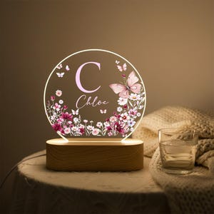 May include: A personalized night light with a wooden base. The clear acrylic light features a floral and butterfly design with the name "Chloe" and the letter "C" in pink. The light is on a round shape.