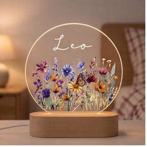 May include: A circular acrylic night light with a warm glowing edge, featuring a printed design of colorful wildflowers and a monarch butterfly. The name Leo is written in white script at the top. The light sits on a smooth, oval wooden base.