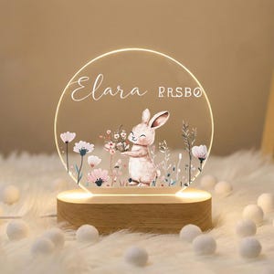 May include: A decorative night light featuring a circular acrylic panel with a bunny illustration holding flowers. The panel is illuminated by a warm light and sits on a wooden base. The name "Elara" is written on the panel.