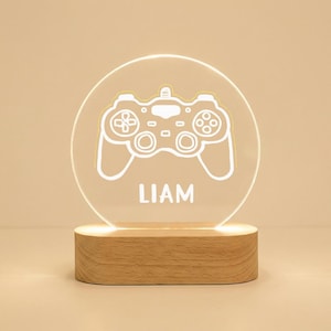 Könnte beinhalten: Ein Nachtlicht zum Thema Gaming mit einer weißen Umrisszeichnung eines Gamecontrollers auf einer transparenten, kreisförmigen Acrylplatte. Der Name "LIAM" ist darunter aufgedruckt. Die Platte steht auf einem rechteckigen Holzsockel.