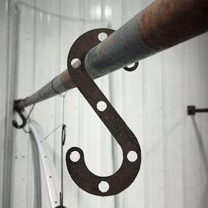 Könnte beinhalten: Ein rostiger, metallener S-förmiger Haken mit kreisförmigen Löchern, der an einem verwitterten Metallrohr befestigt ist. Das Rohr ist an einer weißen Wand montiert. Zusätzliche Haken und ein gebogenes weißes Objekt hängen darunter.