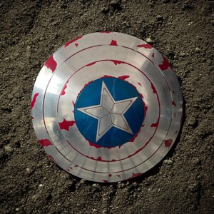 May include: A silver and red shield with a blue center featuring a white star. The shield has a circular design with concentric rings and red accents around the edges, resting on a textured surface.