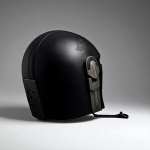 May include: A matte black helmet with a silver-colored visor and accents. The helmet has a rounded top and a detailed, angular design on the lower back. A black strap hangs from the side. The helmet is likely for cosplay or costume use.