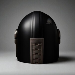May include: A matte black helmet with a vertical stripe down the center. The helmet has a brown leather-like chin strap and side accents. A small emblem is visible on the upper right side of the helmet.