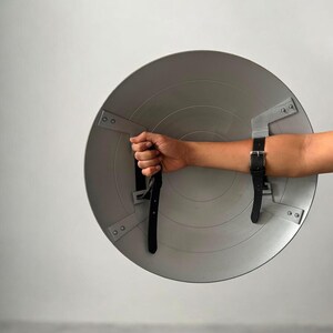May include: A silver-colored, round shield with black leather straps and buckles. The shield is held by a person's hand and forearm. The shield has a metallic appearance and is designed with a series of geometric shapes.