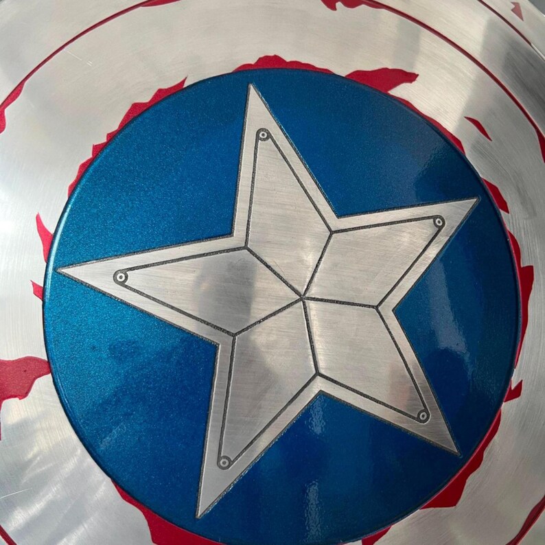 May include: A silver shield with a blue central circle featuring a large, five-pointed star. The star is silver with a detailed outline. The shield has a red border and a metallic finish, reminiscent of a superhero's iconic accessory.