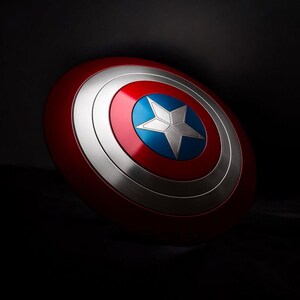 May include: A red, white, and blue shield with a silver star at its centre. The shield has a circular design with concentric rings of red, silver, and blue. It is a prop or collectible item.