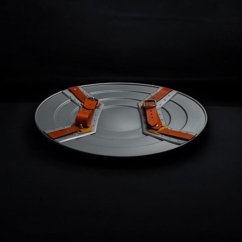 May include: A silver-coloured, circular shield with two brown leather straps and metal accents. The shield has a central, raised circular design and is set against a black background. The straps have metal buckles.