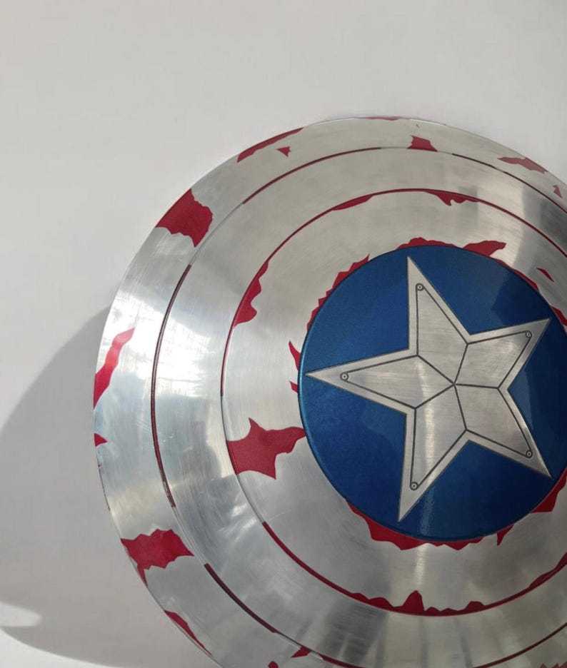 May include: A silver shield with a blue center and a white star. The shield has red accents around the edges. The shield is a replica of Captain America's shield.