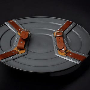 May include: A grey, circular shield with brown leather straps and silver metal detailing. The shield features rivets and a buckle on each strap. This is a prop or collectible item.