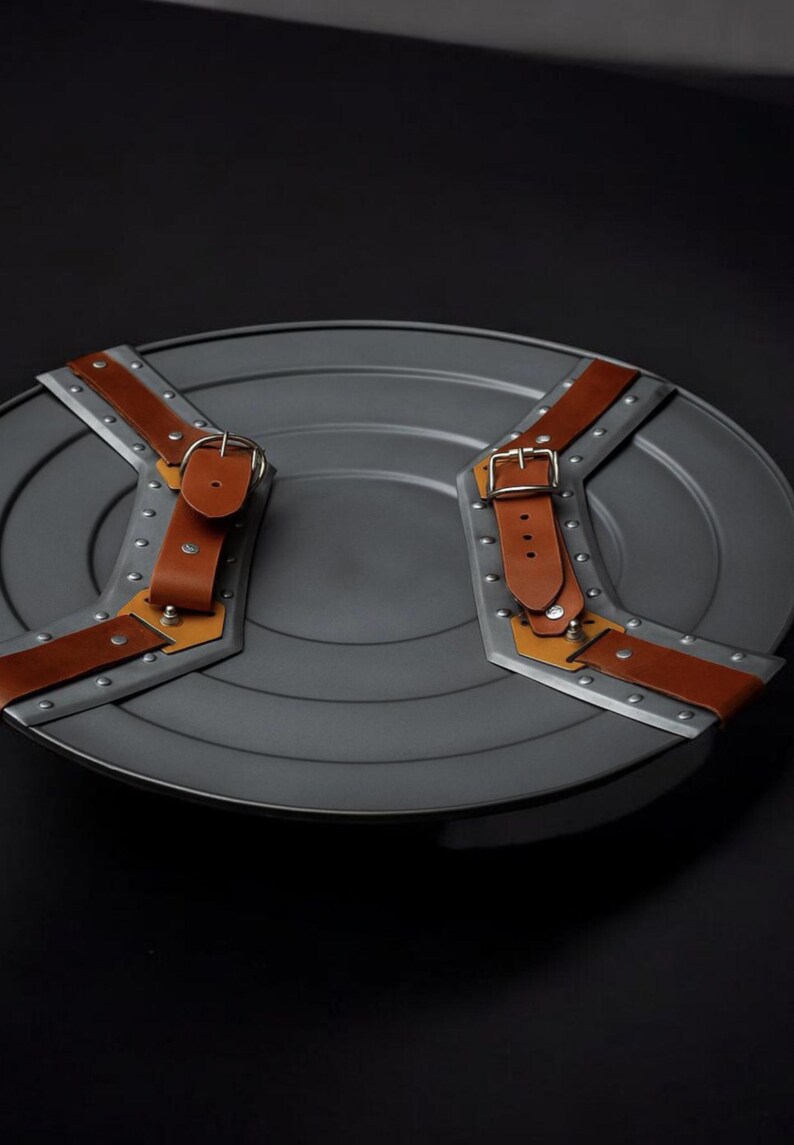May include: A grey, circular shield with brown leather straps and silver metal detailing. The shield features rivets and a buckle on each strap. This is a prop or collectible item.