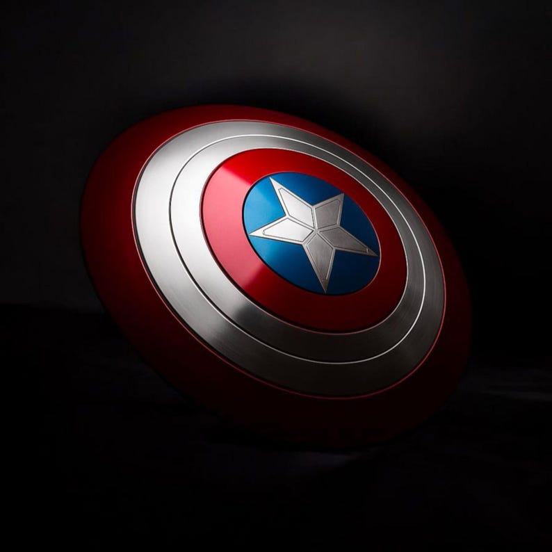 May include: A red, white, and blue shield with a silver star in the center. The shield has a circular design with concentric rings of red, silver, and blue. The shield is a prop or collectible.