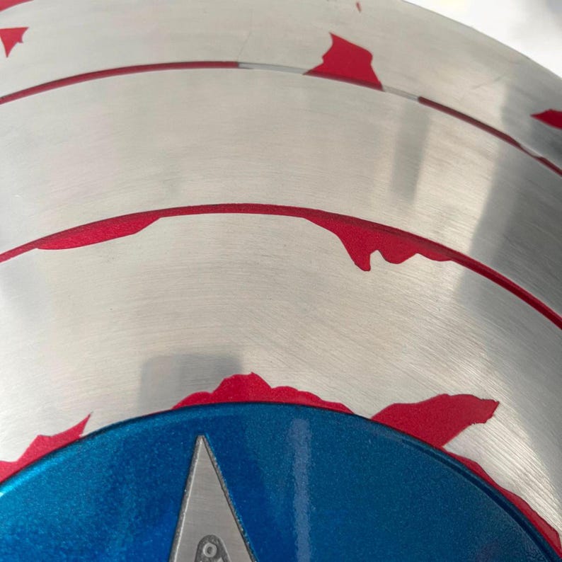 May include: A close-up of a silver shield with red accents and a blue central section. The shield features a silver star in the center. The red accents are around the edge of the shield.