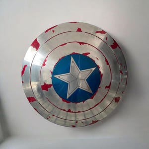 May include: A silver-colored shield with a blue central circle featuring a white star. The shield has red accents around the edges and concentric circles. A superhero-themed collectible.
