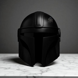 May include: A matte black Mandalorian helmet with a distinctive T-shaped visor and a crest detail on the crown. The helmet is displayed on a white marble surface, showcasing its sleek design and iconic shape.