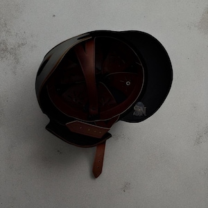 以下が含まれることがあります： 茶色の革の内装とあご紐が付いた黒いヘルメット。ヘルメットはバイザー付きの丸みを帯びた形状で、耐久性のある素材で作られている可能性があります。内部には、しっかりとフィットするように革製のストラップが付いています。