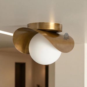 Op de afbeelding: Een moderne plafondlamp met een messing afwerking. De lamp heeft een wit bolvormig licht omgeven door gebogen messing elementen. Het ontwerp is eigentijds, geschikt voor diverse interieurstijlen.