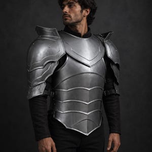 Könnte beinhalten: Ein silberfarbener Brustpanzer und Schulterpanzer-Set. Die Rüstung hat ein geschichtetes Design mit gebogenen Kanten und wird mit Riemen befestigt. Die Person auf dem Bild trägt ein schwarzes Langarmshirt und eine schwarze Hose.