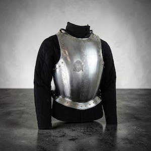 May include: A polished steel breastplate with decorative rivets and etched designs. The armor is displayed over a black quilted garment, suggesting historical or theatrical use. A crest is visible on the breastplate.