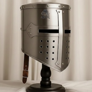 Op de afbeelding: Een zilverkleurige helm in middeleeuwse stijl met een kruisvormig vizier en klinknageldetails. De helm heeft een leren kinriem en wordt tentoongesteld op een zwarte houten standaard. Een wapen is zichtbaar aan de voorkant.