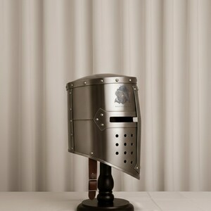 以下が含まれることがあります： 銀色の騎士のヘルメット。バイザーとリベットのディテールがあり、黒い木製のスタンドに展示されています。ヘルメットには紋章と革ストラップが付いています。背景はニュートラルなトーンのカーテンです。