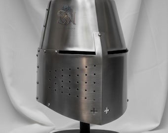 Pode incluir: Um capacete medieval prateado com a parte superior arredondada e a frente plana e angulada. O capacete tem uma série de pequenos orifícios e um recorte em forma de cruz na frente. É exibido em um suporte preto sobre um fundo branco. O capacete tem o texto "SN" e outro texto.
